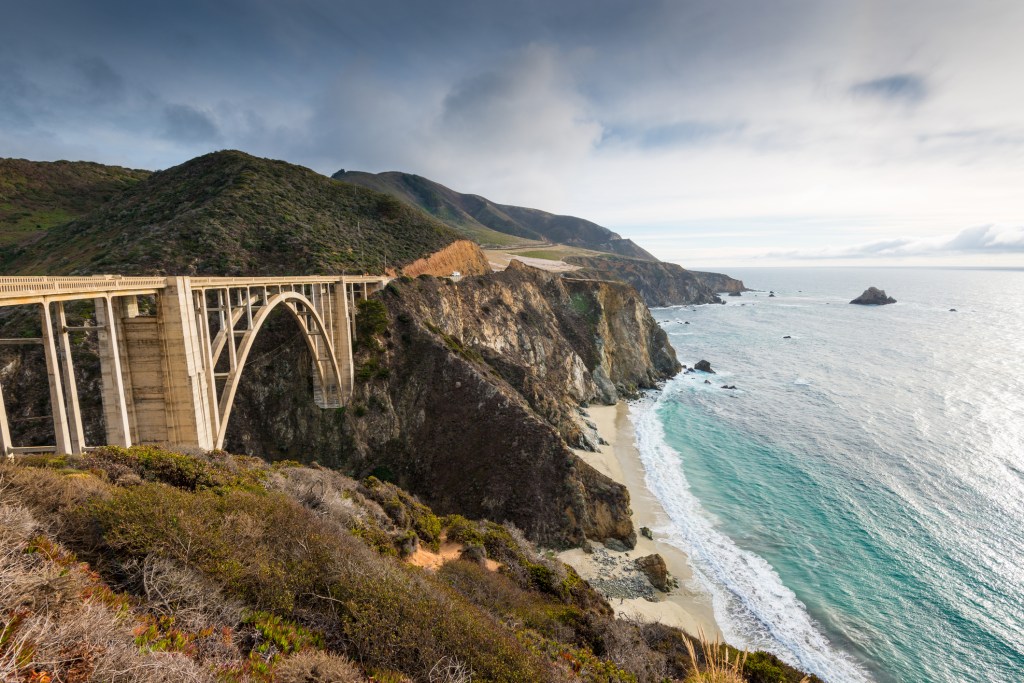 An arched bridge along tall coastal cliffs towering above a long stretch of ocean beach